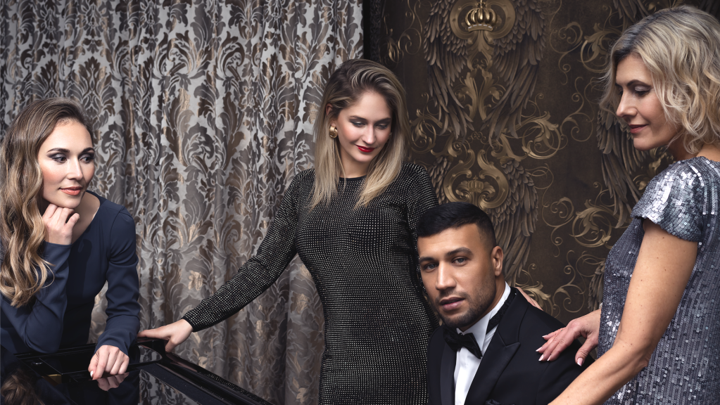 Vier elegant gekleidete Personen, bestehend aus drei Frauen und einem Mann, posieren vor einem gemusterten, dunklen Hintergrund.
